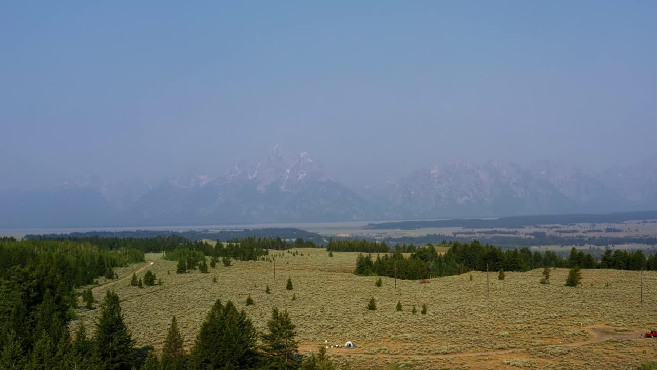 naturaleza de paisaje de drones aéreos que se inclina hacia arriba de la cordillera del parque nacional grand tetons con un valle de matorrales y pinos y un pequeño campamento debajo en un cálido día de verano brumoso en wyoming