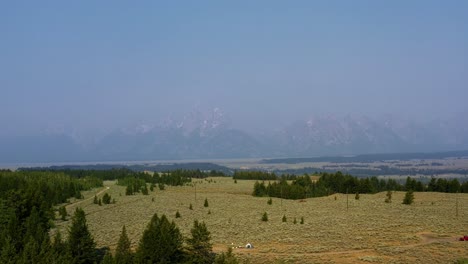 aerial drone landscape nature tilting up shot of the grand tetons national park mountain range with a valley of brush and pine trees and small campground below on a warm hazy summer day in wyoming