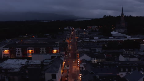 Backwards-reveal-of-straight-illuminated-street-in-evening-town.-Overcast-sky-and-lake-in-distance.-Killarney,-Ireland