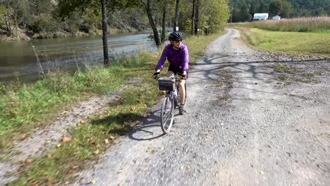 Hübsche,-Reife-Frau-Auf-Einem-Mountainbike,-Die-Auf-Einer-Schotterstraße-Mit-Einem-Fluss-Auf-Der-Einen-Seite-Und-Ackerland-Auf-Der-Anderen-Seite-Fährt