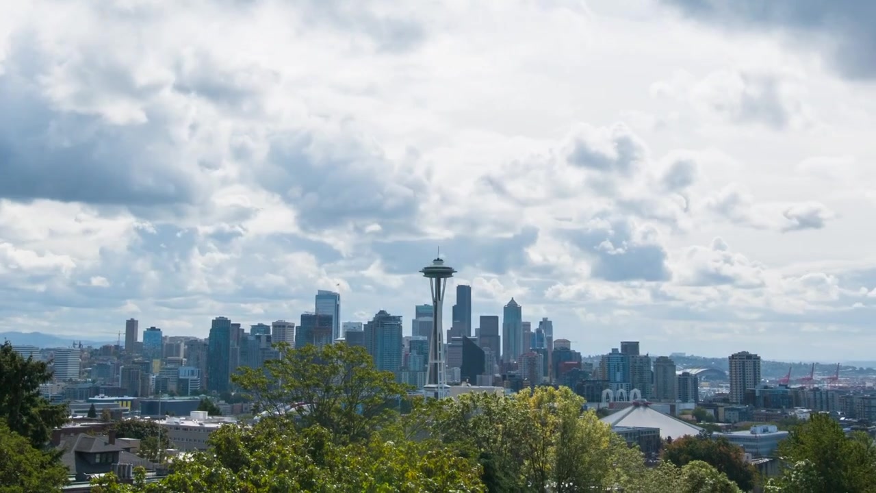 Time lapse che mostra lo Space Needle e lo skyline del centro di Seattle mentre grandi nuvole scorrono nel cielo blu. Gli alberi incorniciano il primo piano con i grattacieli sottostanti