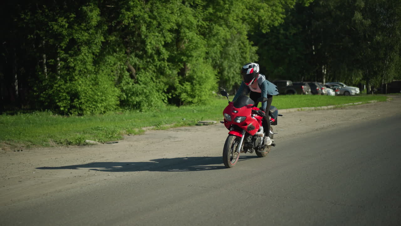 una dama monta una bicicleta eléctrica roja, de pie hábilmente mientras se equilibra en movimiento, su camisa flota en el viento, revelando parcialmente su cuerpo, navegando en un día soleado a lo largo de un camino despejado