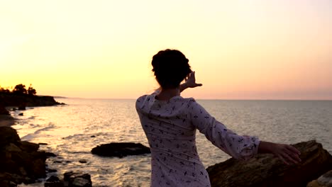 adorable chica caucásica morena en vestido claro bailando en el acantilado sobre el mar a la hora del amanecer