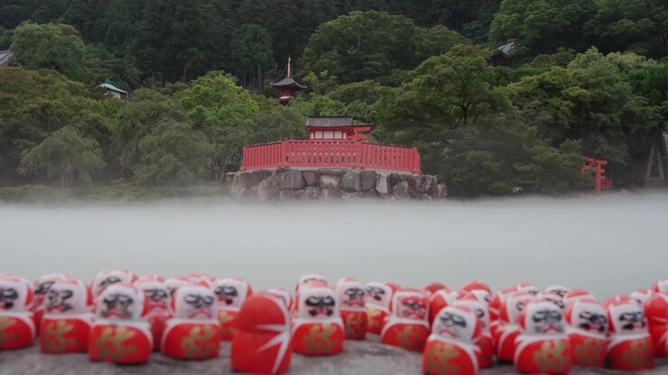 Molte bambole Daruma rosse collocate su una piattaforma di pietra al Tempio Katsuoji a Osaka, circondate da una lussureggiante vegetazione verde e una leggera nebbia, con una struttura tradizionale di tempio rosso visibile sullo sfondo.