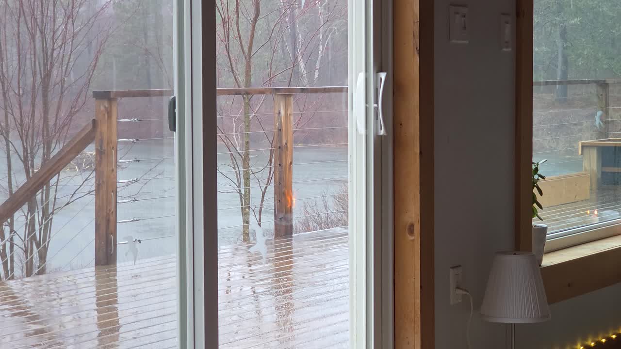 Fireplace reflects in cabin window as rain falls on terrace by forest lake