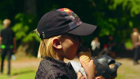 experimentar el deleite de un niño rubio despreocupado de 7 años con cabello largo, poniéndose una gorra, saboreando un cono de helado en un parque o bosque soleado