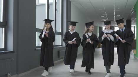 group of happy multiracial kindergarten students in cap and gown walking in the school corridor while holding diploma and celebrating their graduation