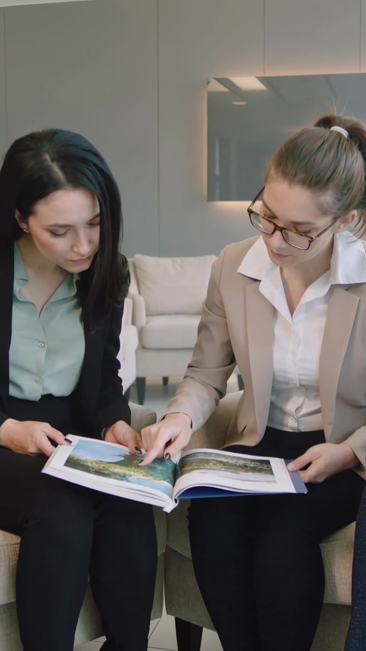 Video verticale: Una donna a destra con un blazer beige indica un catalogo, guidando una collega nella lounge dell'ufficio.