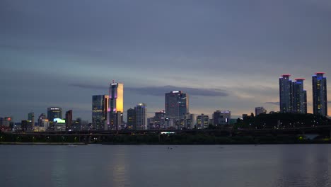 night seoul from the riverbank of han river on colorful sunset, landscape of skyscrapers like glass tower, trade tower, and asem tower under sunlight, yacht pass by when people paddleboarding on river