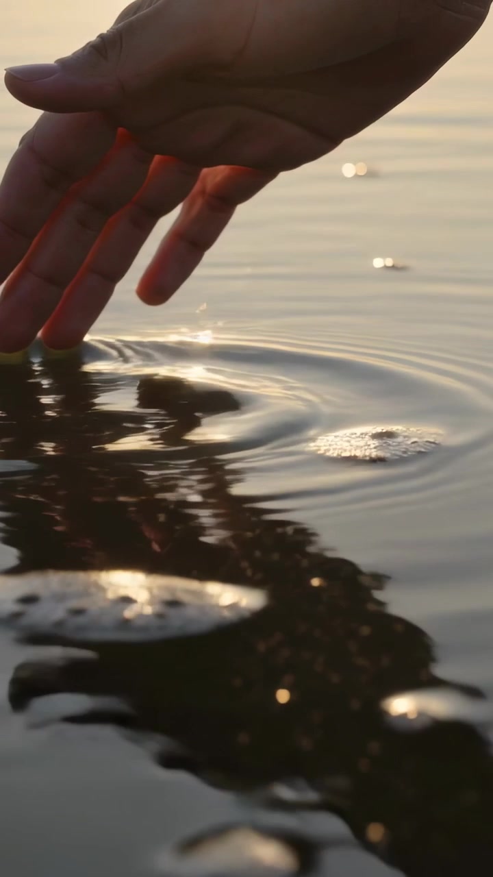 Video verticale: Mano adulta che si avvicina sfiorando l'acqua bassa di un lago, creando increspature e riflessi del sole.