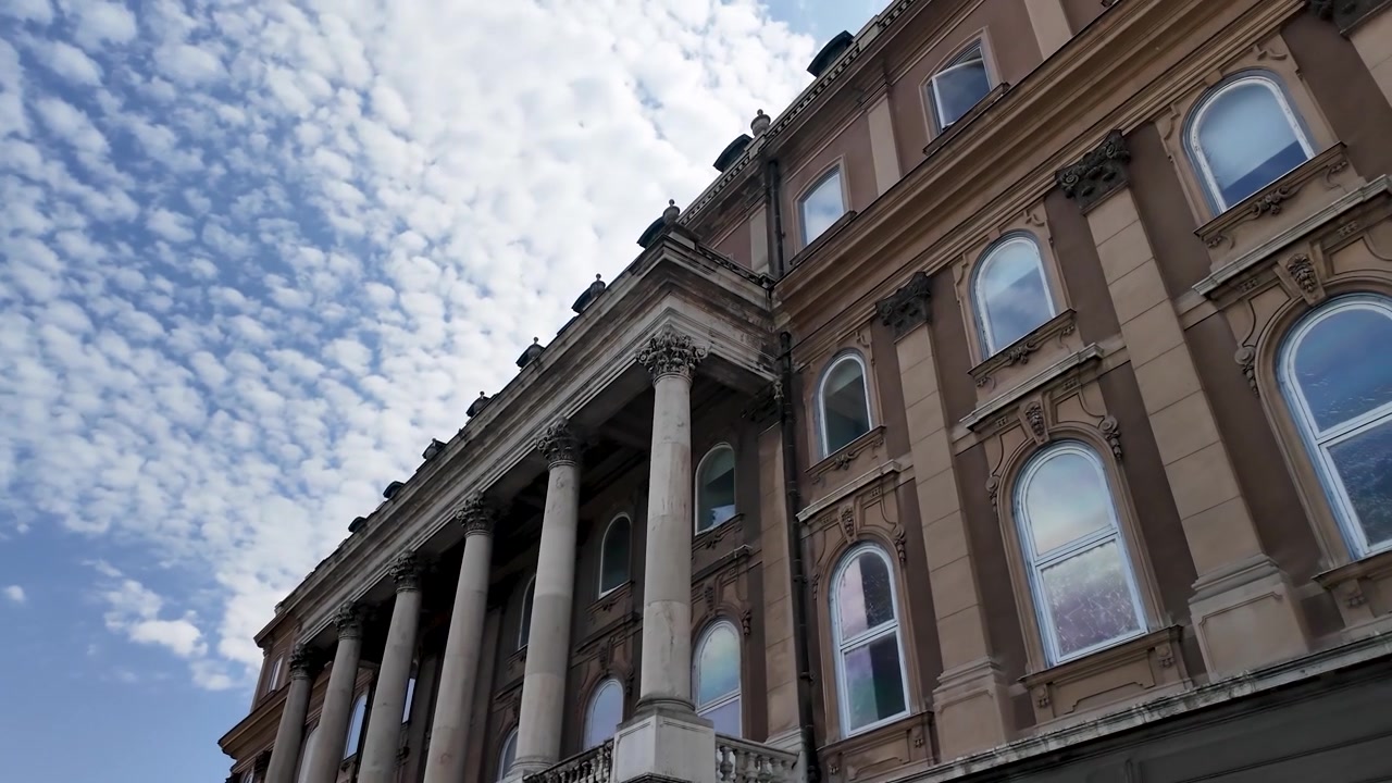 Panoramica cinematografica sulla grande facciata del Castello di Buda a Budapest, Ungheria, che mette in evidenza colonne neoclassiche, architettura storica e cieli drammatici pieni di nuvole.