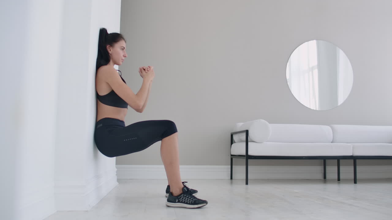 The brunette girl froze in a squat leaning on the wall doing a static exercise in a bright interior. Keep 90 degree leg angle