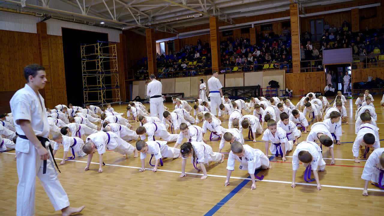 Young karate athletes doing push-ups in the huge gym. Coaches watch the sportsmen performing exercise. Spectators at the background.