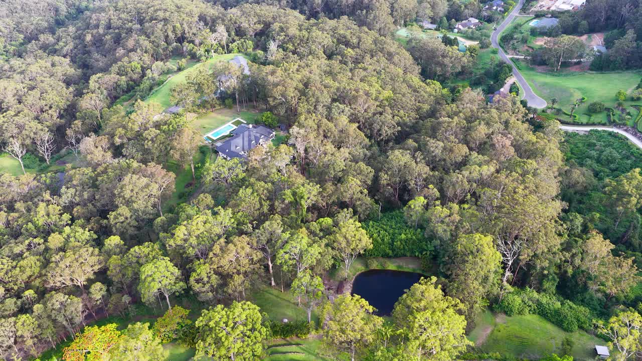 Drone glides over forested luxury estate, revealing house, pool, pond, and lush greenery