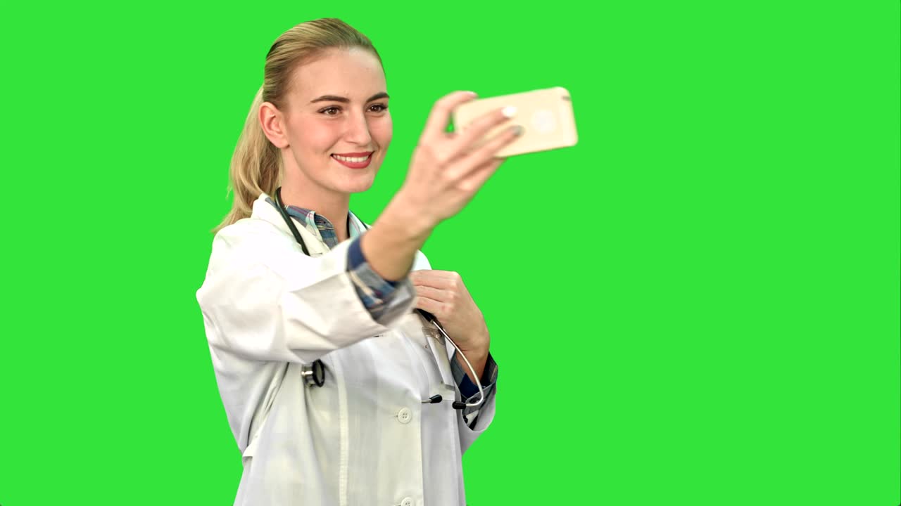 젊은 예쁜 여성 의사는 녹색 화면, 크로마 키에서 스마트 폰으로 셀피를 만니다.