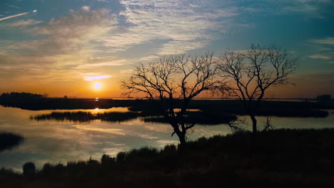 aerial shot during a peaceful, colorful sunset, moving in toward two dead trees at the edge of a glistening lake