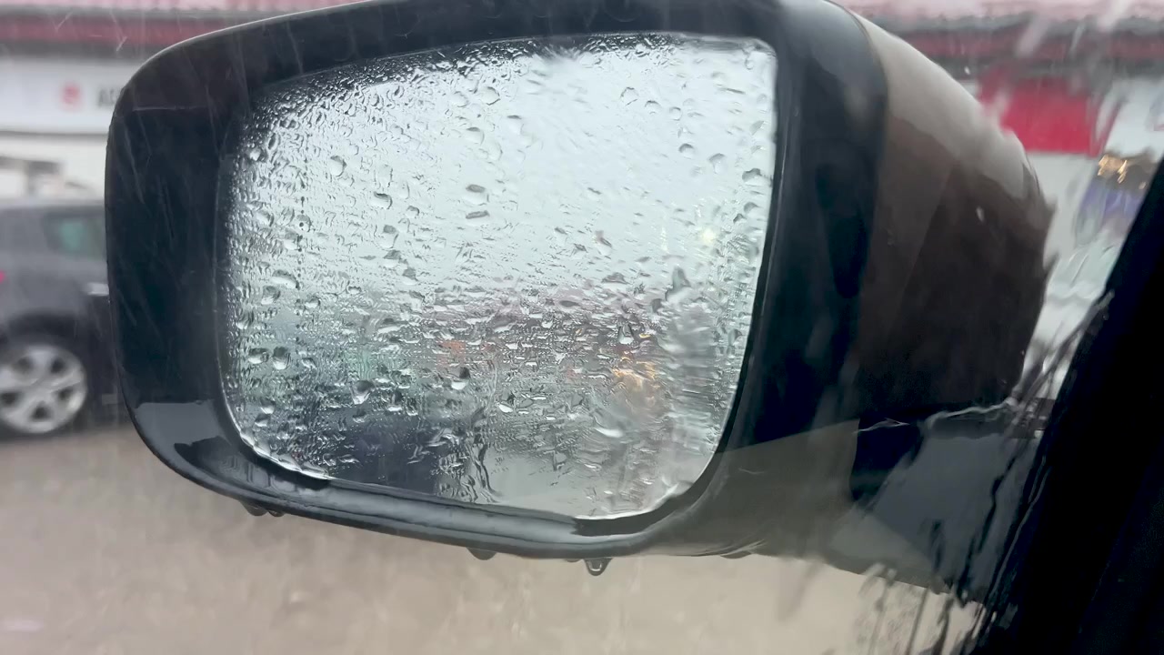 Primo piano di goccioline d'acqua su uno specchietto retrovisore di un veicolo durante una giornata piovosa. Autentica scena meteorologica urbana per concetti di viaggio e sicurezza.