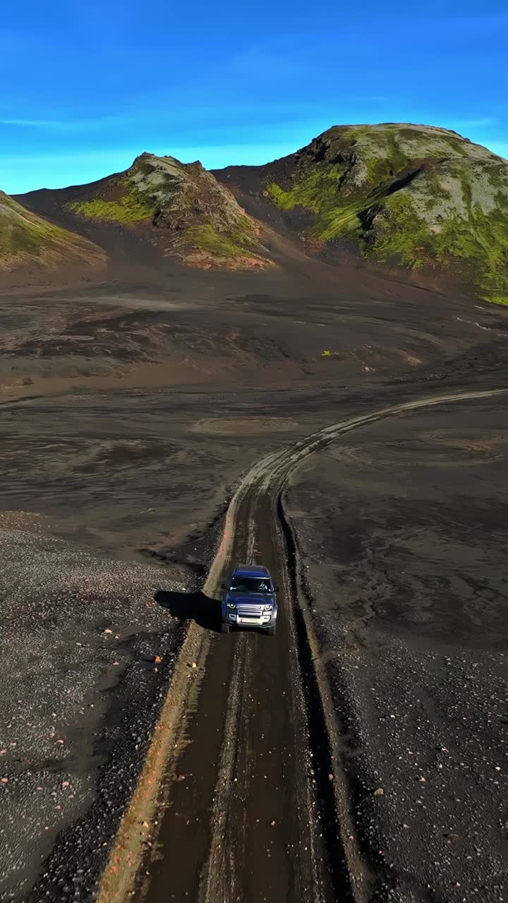 Ripresa verticale con drone che segue un SUV 4x4 mentre percorre un sentiero sterrato attraverso il deserto vulcanico nero degli altopiani islandesi verso montagne ricoperte di muschio sotto un cielo azzurro.