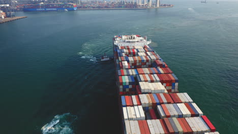 Giant-container-vessel-pushed-towards-the-dock-by-tugboats-for-unloading,-with-the-Stonecutters-bridge-and-container-port-in-the-background-in-Hong-Kong