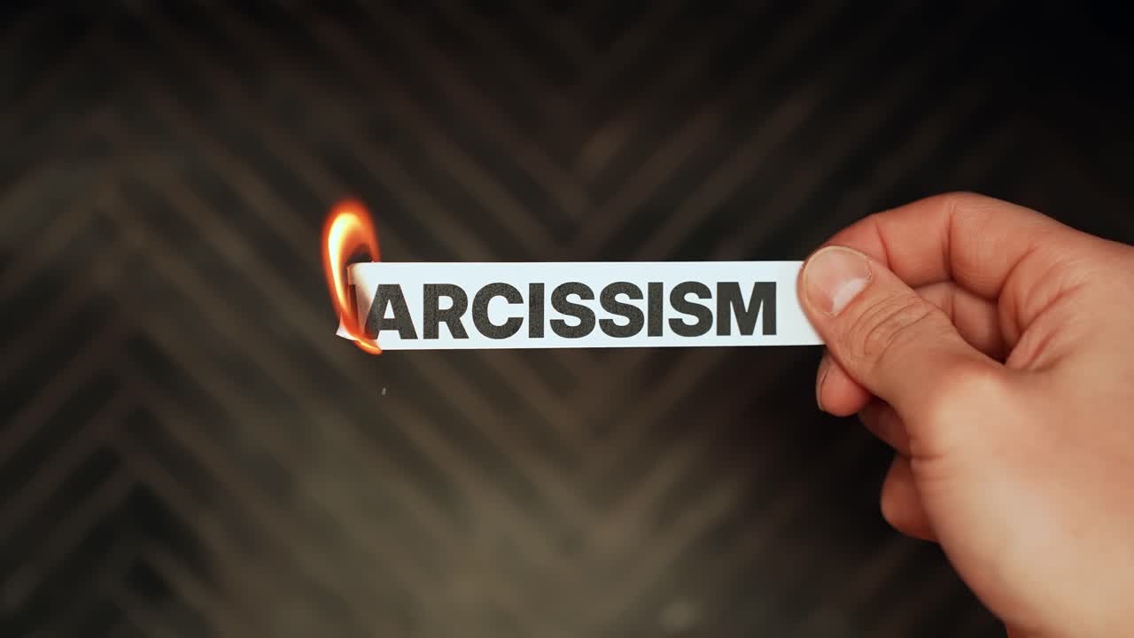 Hand burns paper label reading “NARCISSISM” with flame on dark background, toxic personality and mental health concept, copy space