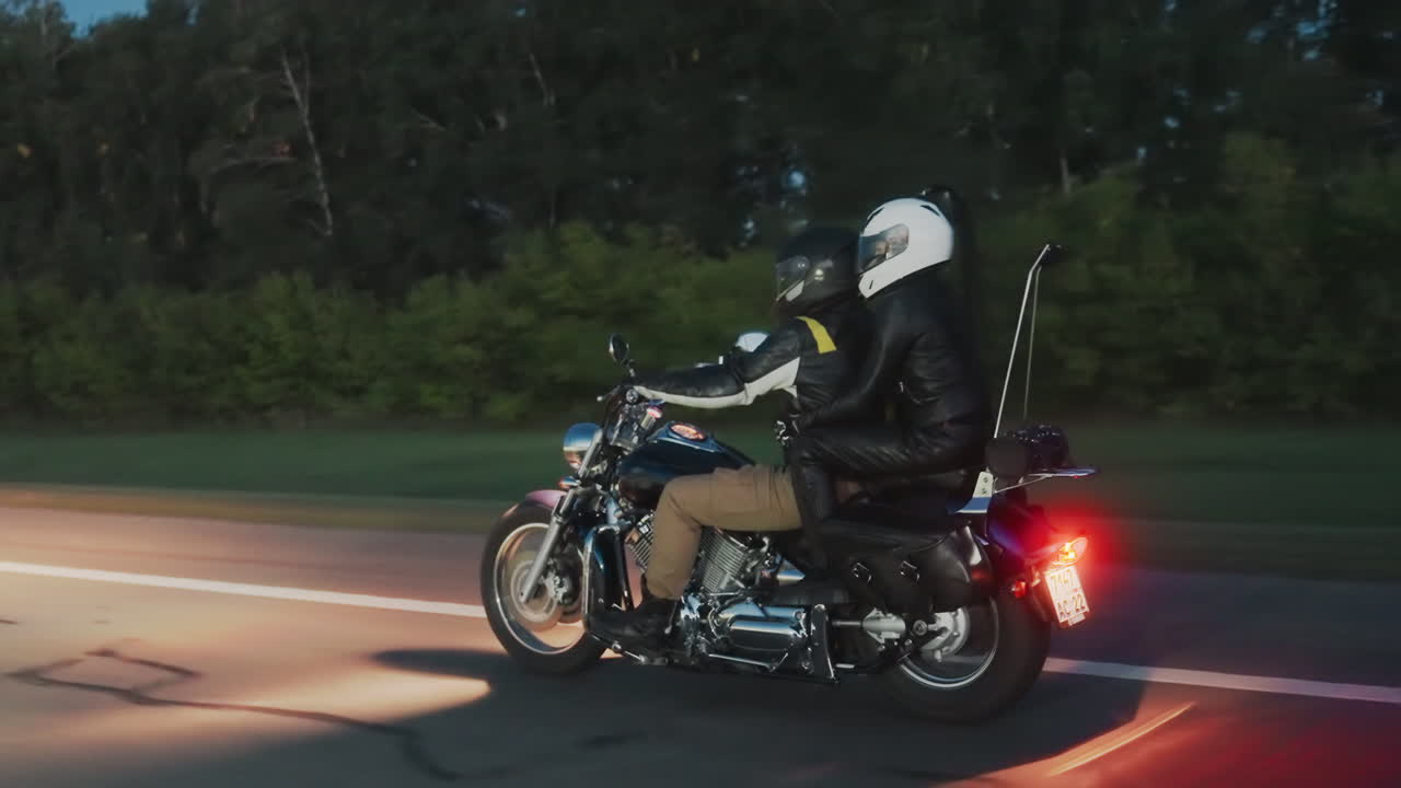 Motorcyclist with passenger rides past signpost on evening highway, both wearing helmets and leather jackets, motorcycle illuminated by road lights, surrounded by trees and smooth asphalt
