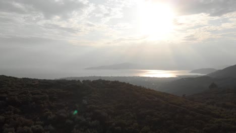 aerial  day drone shot of amazing mountain landscape  green tall trees small traditional village sun shines behind white small clouds sun reflects in the sea at background amazing view aegean sea
