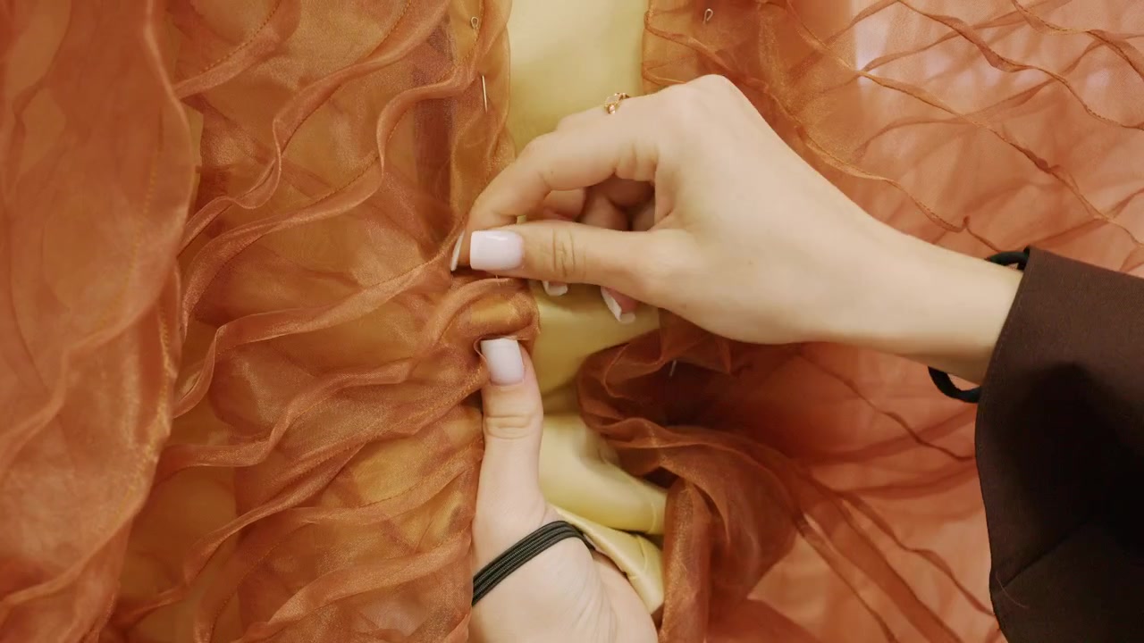 Designer's hands hold a round pin cushion filled with silver dressmaker pins while working on layered orange organza fabric