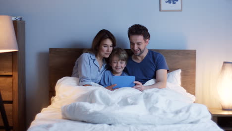 Caucasian-parents-sitting-on-the-bed-with-their-little-cute-son,-watching-something-on-the-tablet-and-talking-at-night