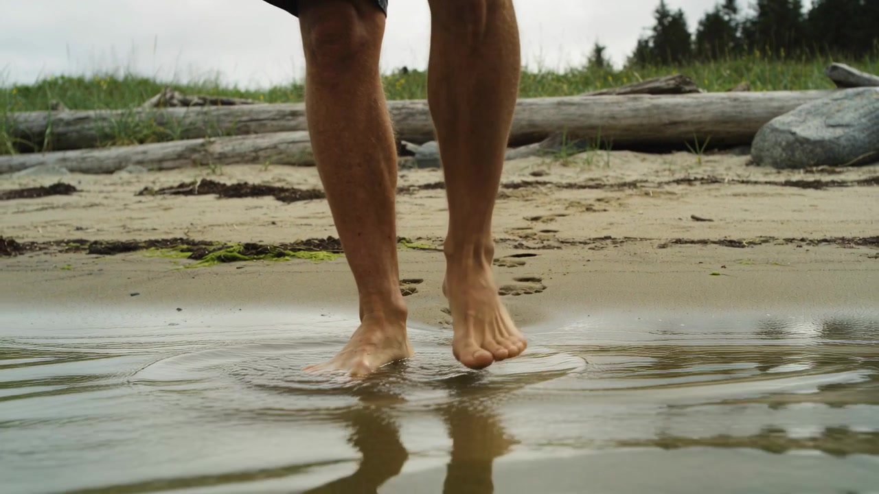 Walking man in dark shorts lifting foot stepping into shallow water at shore leaving wet prints