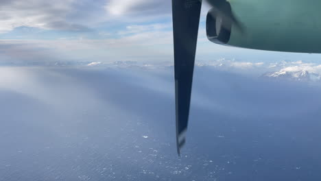 Domestic-Aircraft-Norway,-Turboprop-engine-in-foreground,-shot-out-of-window-in-northern-Norway,-with-snow-capped-mountains,-mostly-cloudy-sky,-and-foggy-ocean-view,-Lofoten-Island,-right-to-left-shot