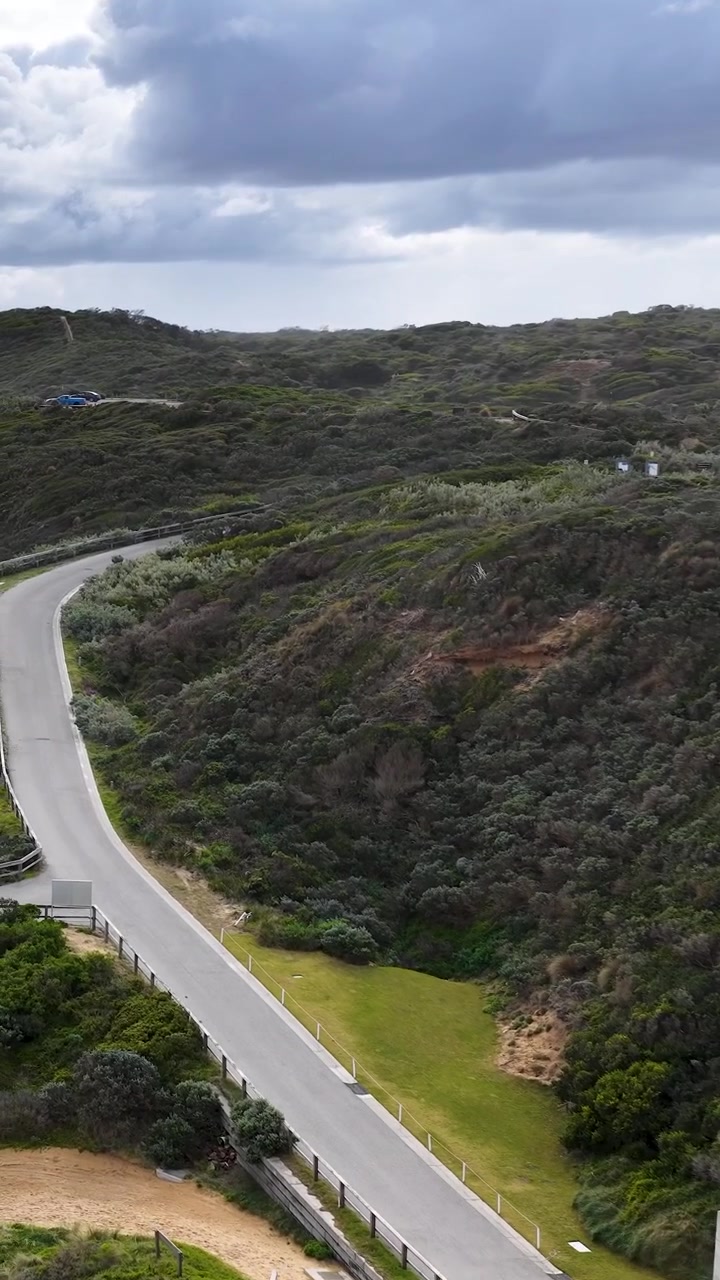 Il drone si muove con una panoramica sopra una strada costiera tortuosa, architettura moderna, colline lussureggianti, sotto un cielo diurno coperto.