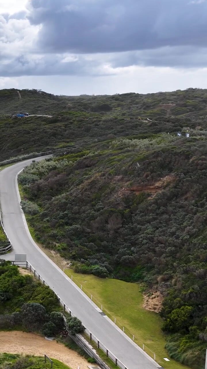 Il drone si muove con una panoramica sopra una strada costiera tortuosa, architettura moderna, colline lussureggianti, sotto un cielo diurno coperto.