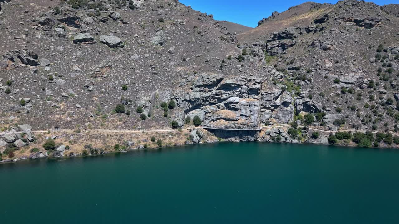 Drone approaches the Dunstan Trail in Central Otago, revealing the winding path beside Lake Dunstan and rugged rocky hills on this famous New Zealand Great Ride