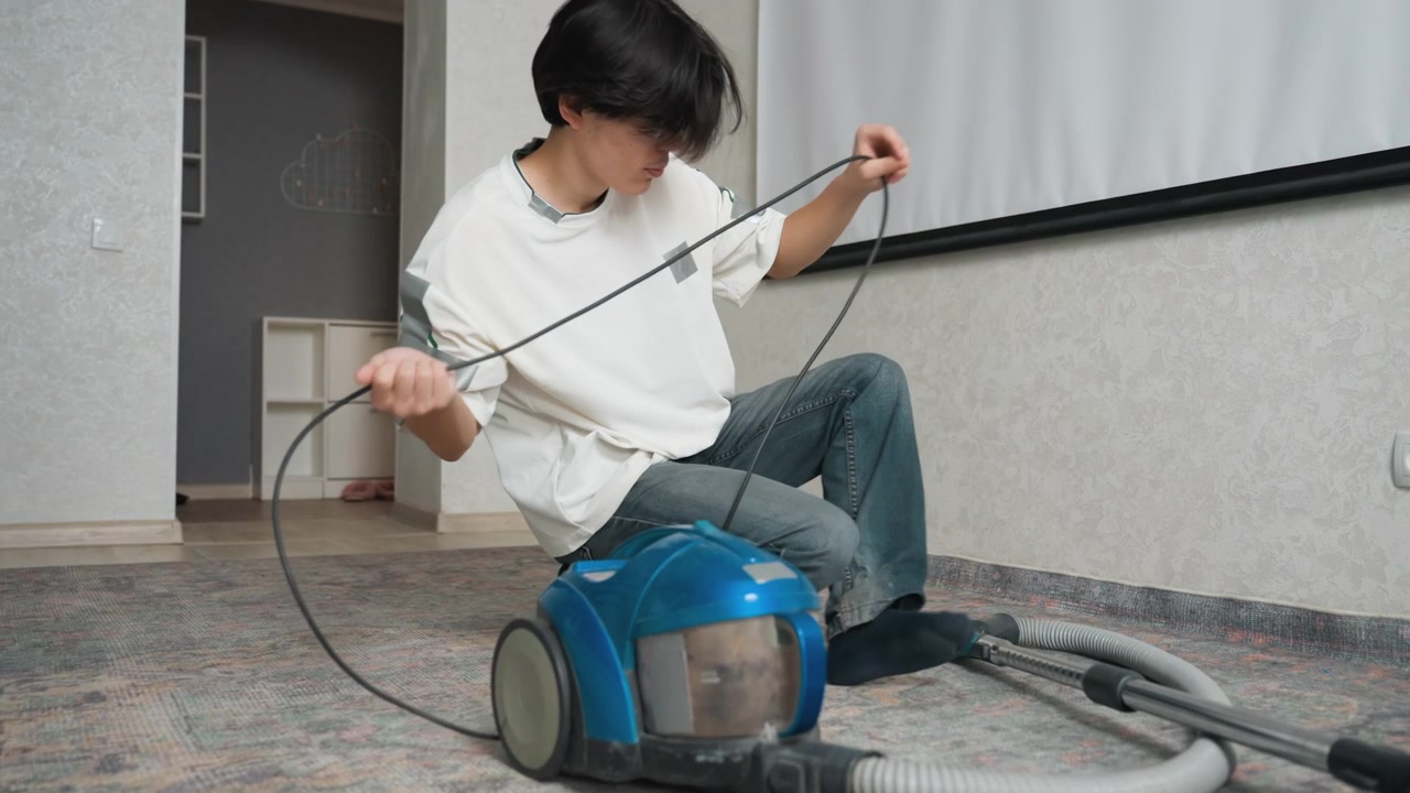 空のリビングルームで掃除機を修理するアジア人青年。柄物のカーペットに膝をつき、コードとキャニスターを確認し、ホースの接続を点検している。集中したDIYメンテナンス、家事としての掃除。