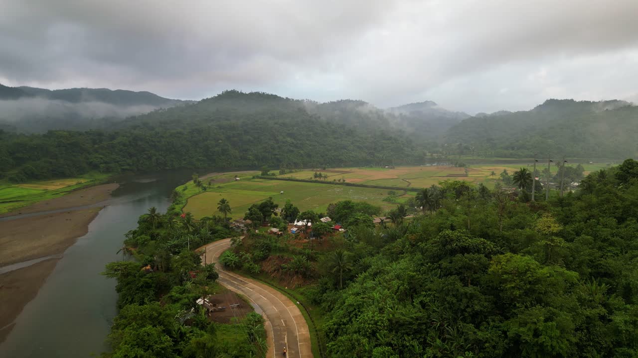 Nuvole nebbiose pendono basse su montagne verdi dietro i campi agricoli e un fiume tropicale vicino alla strada