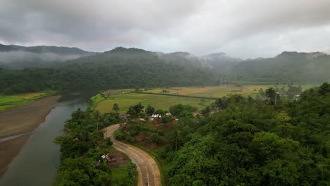 Misty-clouds-hang-low-on-green-mountains-behind-farmland-and-tropical-river-near-road