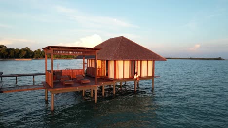 Young-Woman-Enjoys-Sunset-Sitting-On-Edge-of-Over-Water-Bungalow-Resort-at-Leebong-Island,-Belitung-Indonesia---Aerial-Pull-Back