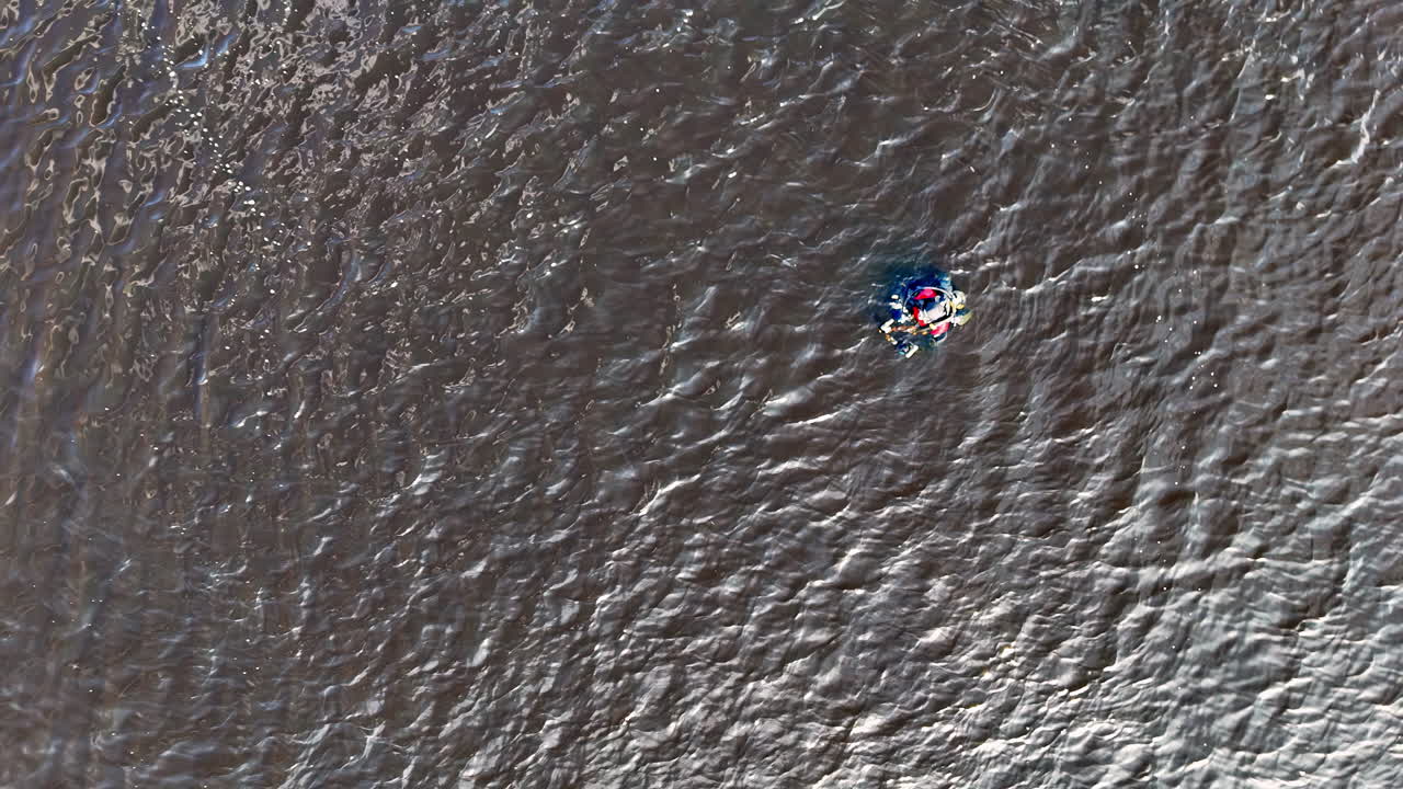 Scuba diver wearing full equipment with visible red and blue accents floats on the surface of rippling water, surrounded by textured waves reflecting shifting light