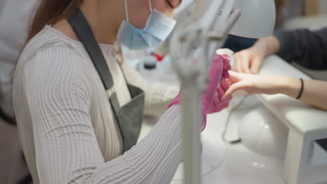nail technician wearing protective face mask and pink gloves carefully grooming customer nails using brush and scissors under bright lamp with blurred metal support in background