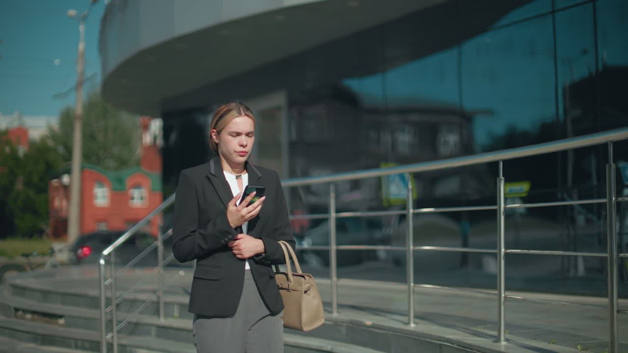 Thoughtful woman in formal attire on phone call walks with focused expression beside glass building reflecting surrounding structures and moving cars in bright urban environment during daytime