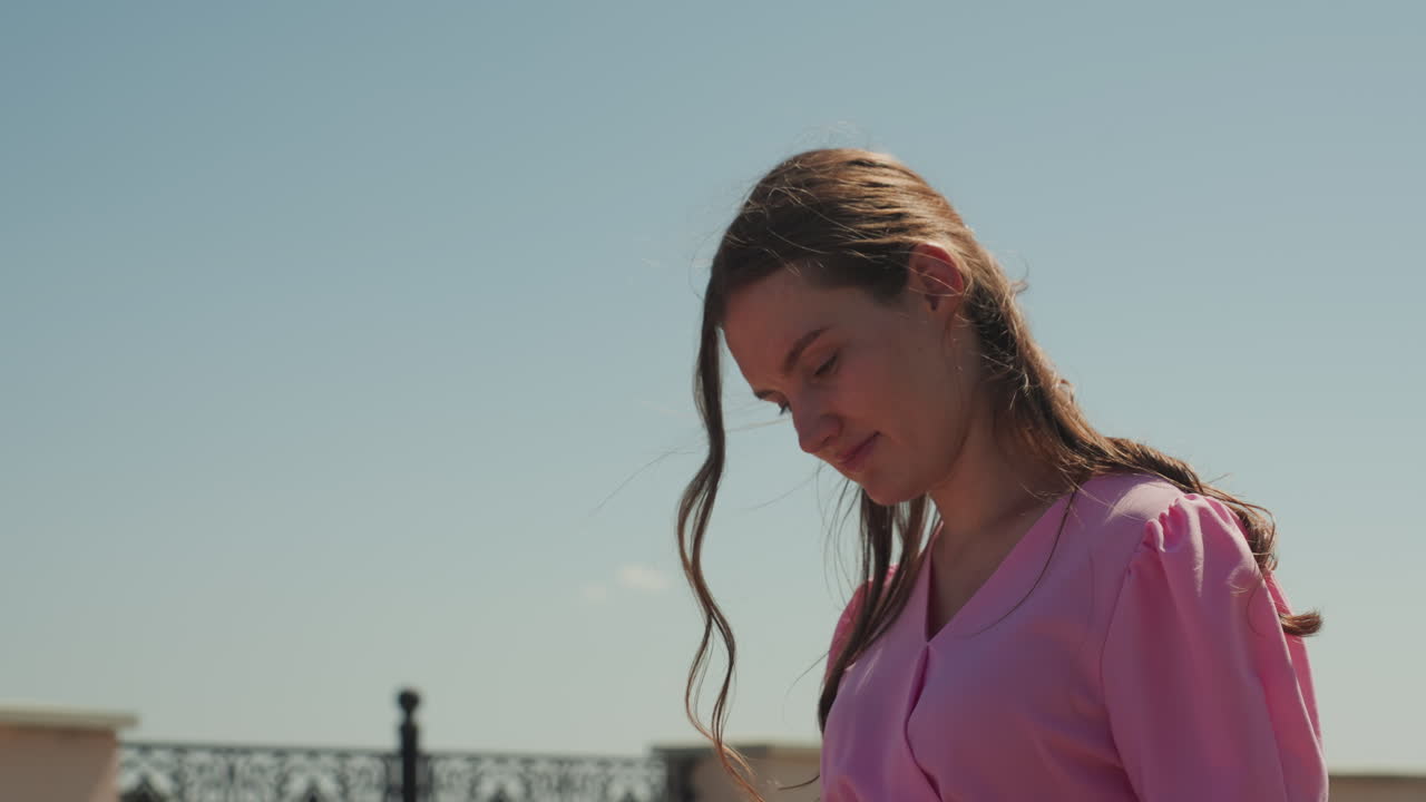 joven caucásica mirando al cielo, perfil seguro con blusa rosa, trenzas ondeando al viento, farola y barandilla ornamentada al fondo, cielo despejado y brillante, tono cinematográfico optimista, optimismo reflexivo
