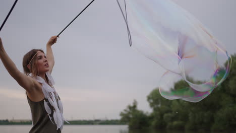 A-young-hippie-woman-in-a-dress-and-with-feathers-on-her-head-makes-huge-soap-bubbles-at-sunset-on-the-shore-of-a-lake-in-slow-motion