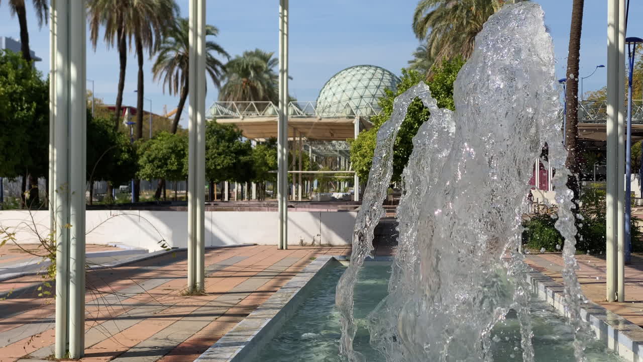 Primer plano de los rítmicos chorros de la fuente en el parque de la Expo de Sevilla, captando la brillante luz del sol sobre los árboles y las estructuras modernas