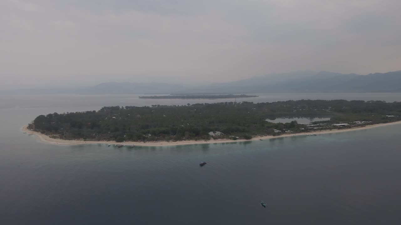 Stunning aerial footage of narrow peninsula surrounded by clear waters, boats, and misty hills near Serangan Island. Captures tranquil scenes from above in soft, diffused lighting