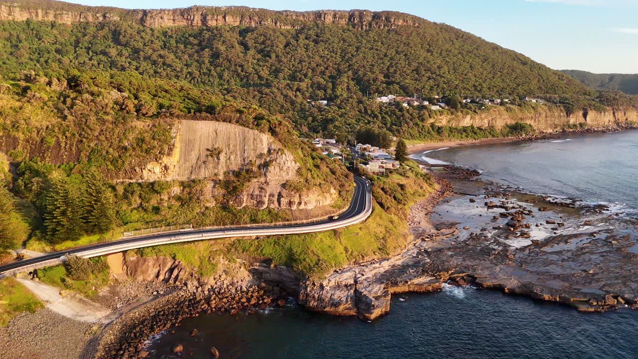 Wide aerial footage of the Sea Cliff Bridge, with a vehicle navigating the curving road above the blue waters of the Pacific. Drone footage.