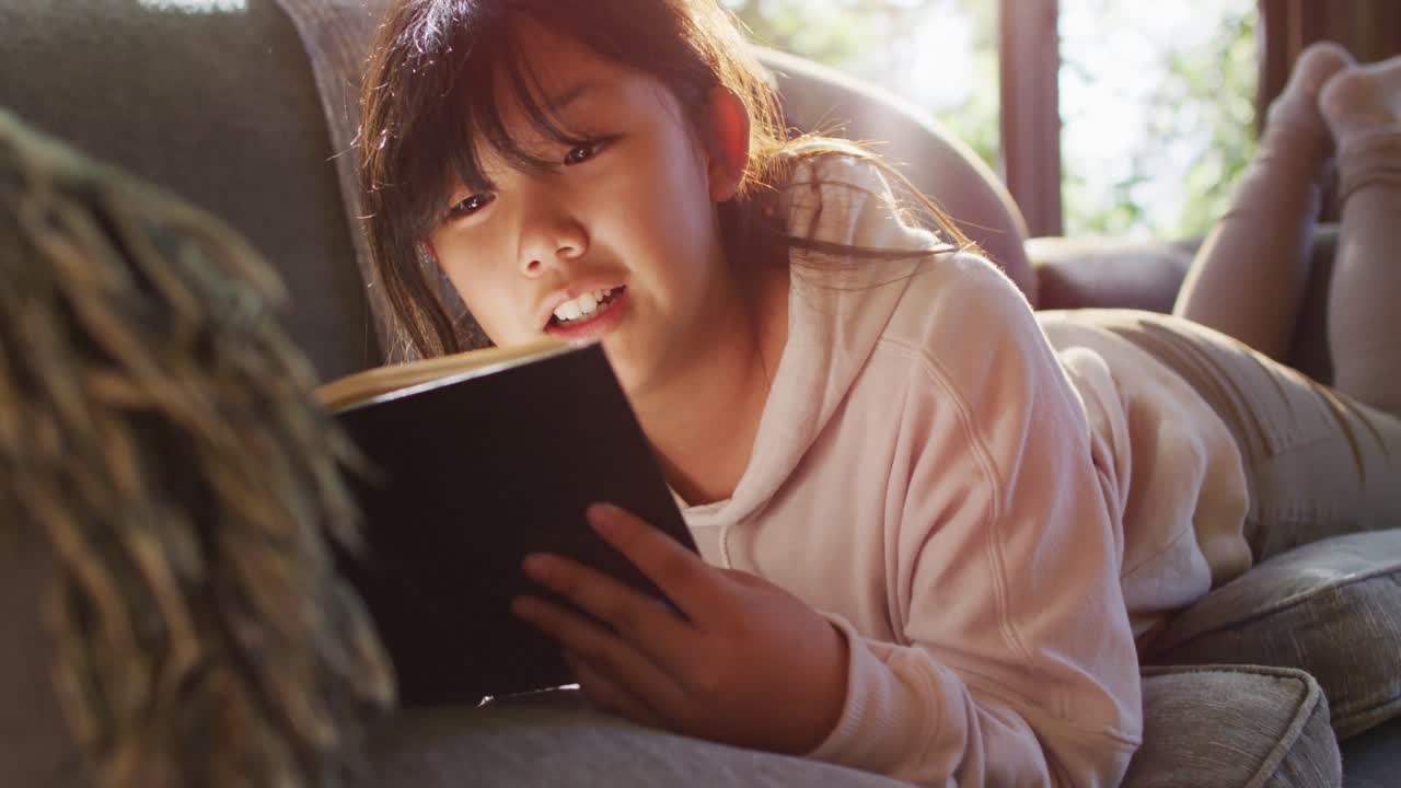 家のソファに寝転がり、笑顔で本を読むアジア系の女の子
