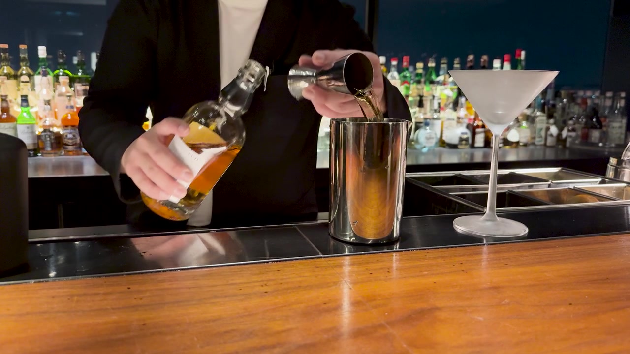 En bartender i sort jakkesæt hælder spiritus i en jigger over en trædisk