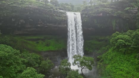 Witness a stunning drone view of Vasudhara Falls in Bhandardara, Maharashtra, India, surrounded by dense forests and lush green mountains. Misty monsoon clouds add a mystical aura