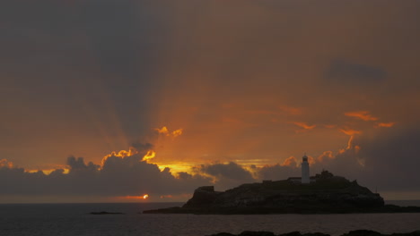 the beautiful sunbeams of the sun setting behind indigo clouds by the godrevy lighthouse in cornwall, england - wide shot