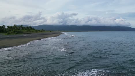 4K-Drohnen-Luftaufnahme-Von-Tosenden-Wellen,-Die-An-Küstenfelsen-An-Einem-Idyllischen-Strand-Mit-Bergen-Und-Bedrohlichen-Wolken-Im-Hintergrund-Treffen